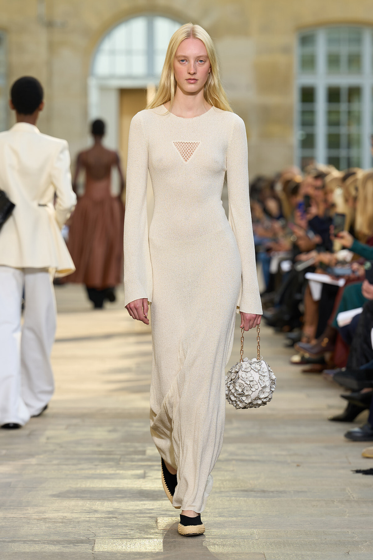 Model walks the runway in a cream knit long-sleeve maxi dress with a small triangle mesh cutout at the chest, holding a textured floral handbag on a chain strap.
