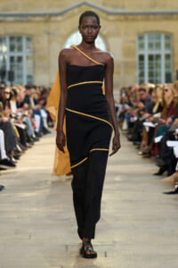 Model walks a runway in a black, one-shoulder dress with gold piping, paired with black sandals; audience watches from both sides in a historic courtyard setting far in the background.