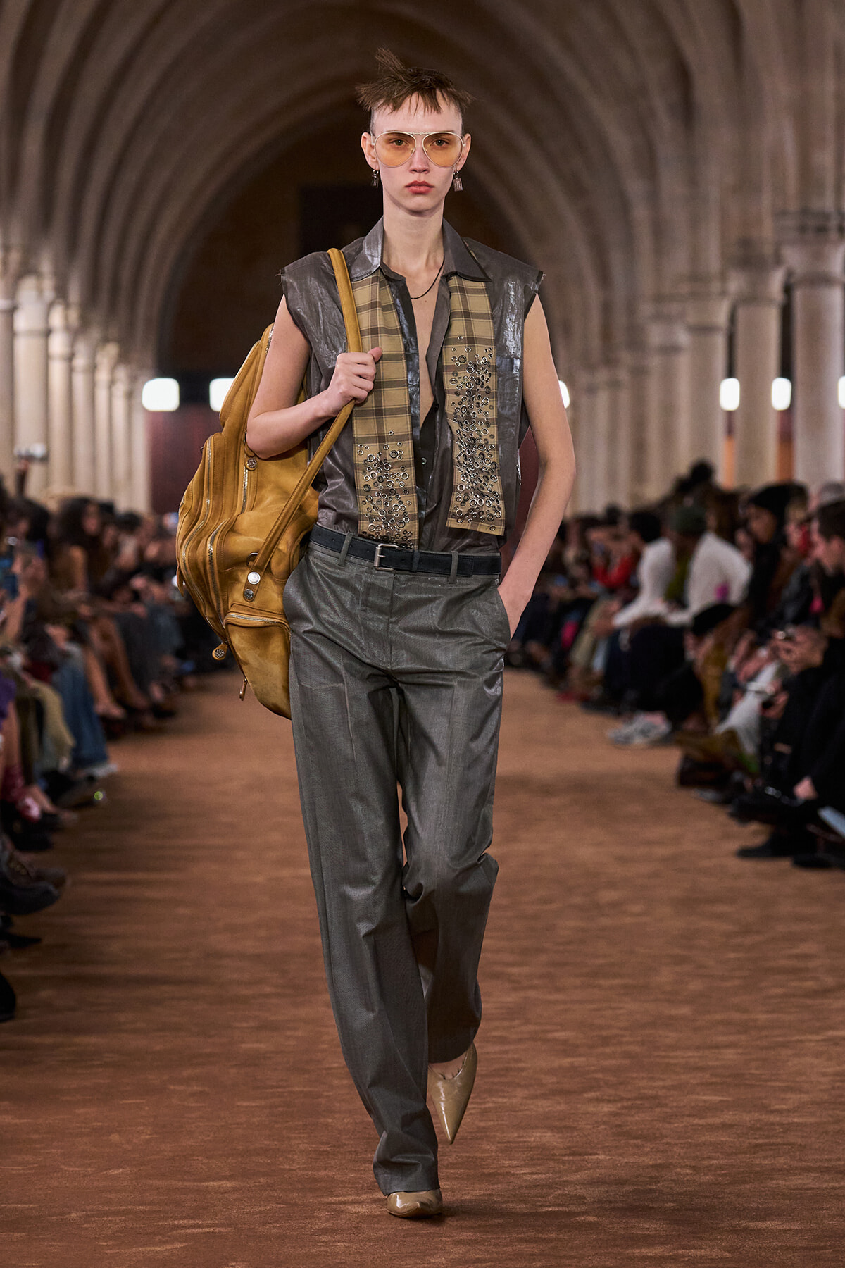 Model walks a runway in a metallic gray sleeveless shirt, embellished plaid vest, gray trousers, and a large tan leather backpack, with amber sunglasses.