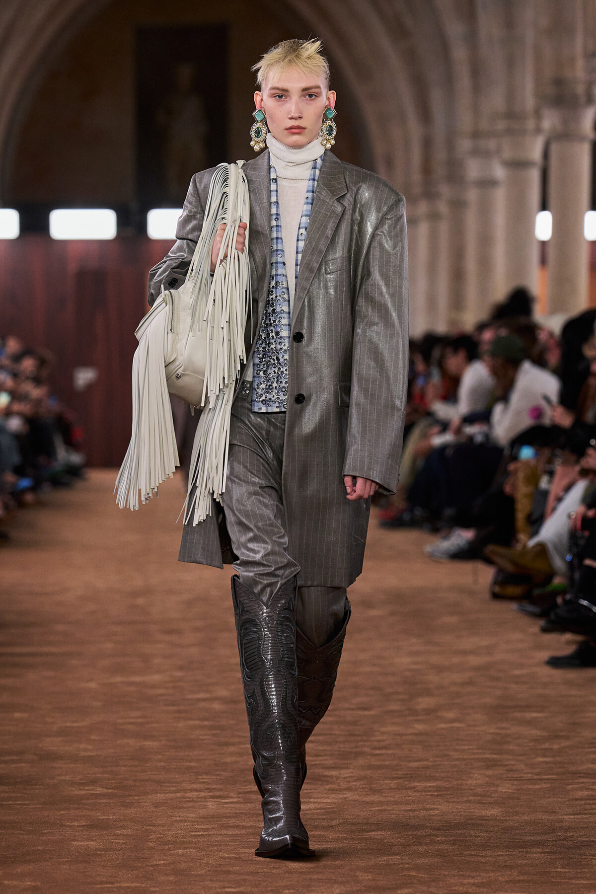 Model on a runway wearing a metallic gray pinstripe suit, white turtleneck, oversized fringe white bag, and tall dark boots, with bold turquoise earrings.