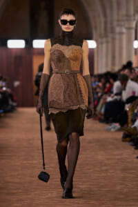 Runway model in a brown lace dress with a belt, fingerless gloves, oversized sunglasses, and a small black handbag on a strap.