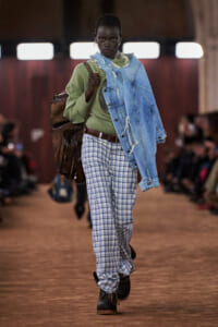 Model walking a runway in a sage-green sweater, oversized distressed denim jacket draped over one shoulder, plaid blue trousers, black boots, and a large brown bag