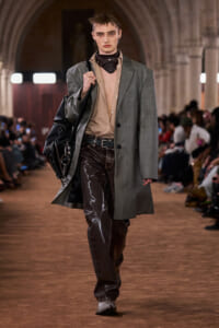 Model walking runway in a gray oversized coat over a beige shirt, shiny dark brown vinyl pants, and a black bag slung over the shoulder.