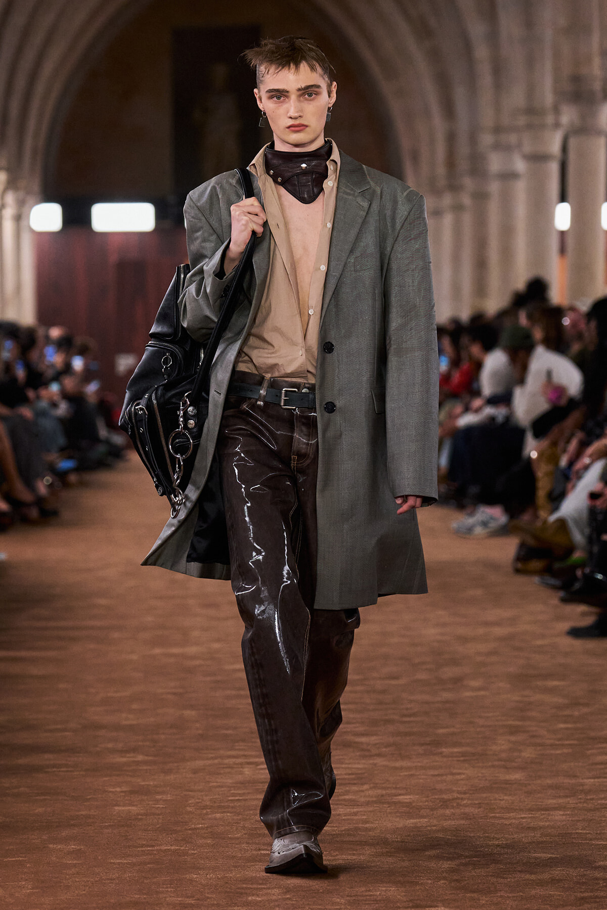 Model walking runway in a gray oversized coat over a beige shirt, shiny dark brown vinyl pants, and a black bag slung over the shoulder.