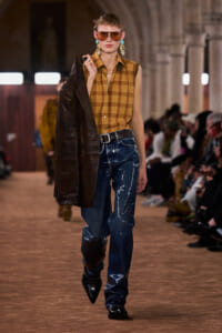 Model on a runway wearing a tan plaid sleeveless shirt, dark jeans with white streaks, and a brown coat draped over the shoulder, oversized sunglasses and turquoise earrings.