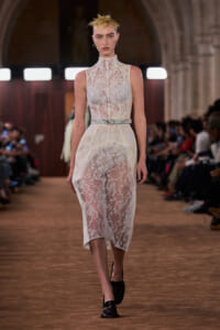 Model walking a runway in a white high-neck lace dress with a sheer overlay and a thin belt.