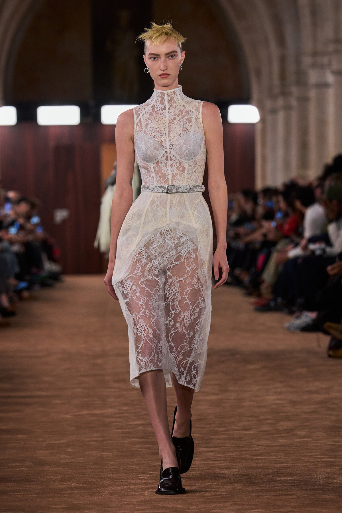 Model walking a runway in a white high-neck lace dress with a sheer overlay and a thin belt.