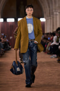 Runway model in a mustard blazer over a blue knit sweater and striped shirt, paired with distressed jeans and a denim bag on a brown floor.