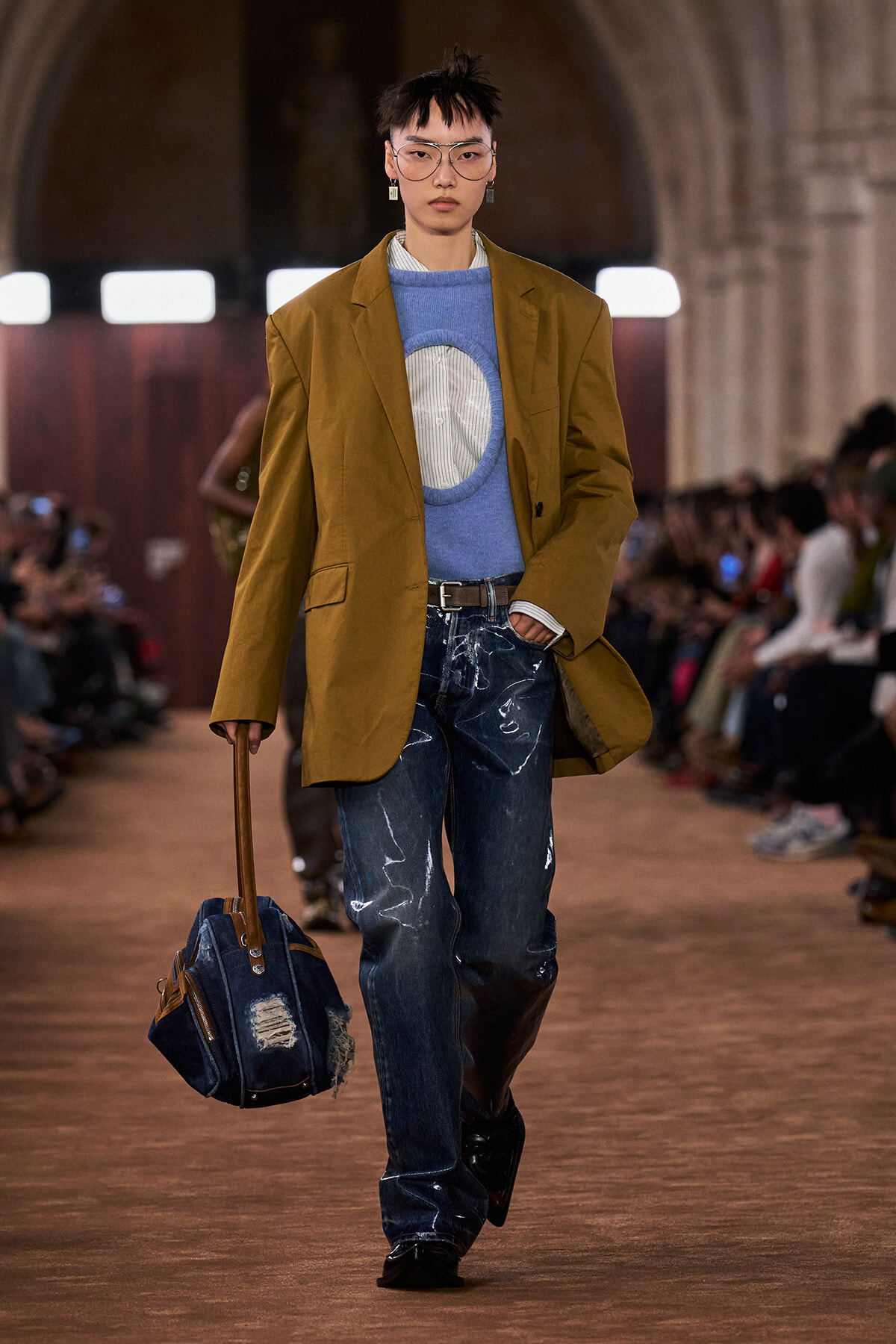 Runway model in a mustard blazer over a blue knit sweater and striped shirt, paired with distressed jeans and a denim bag on a brown floor.