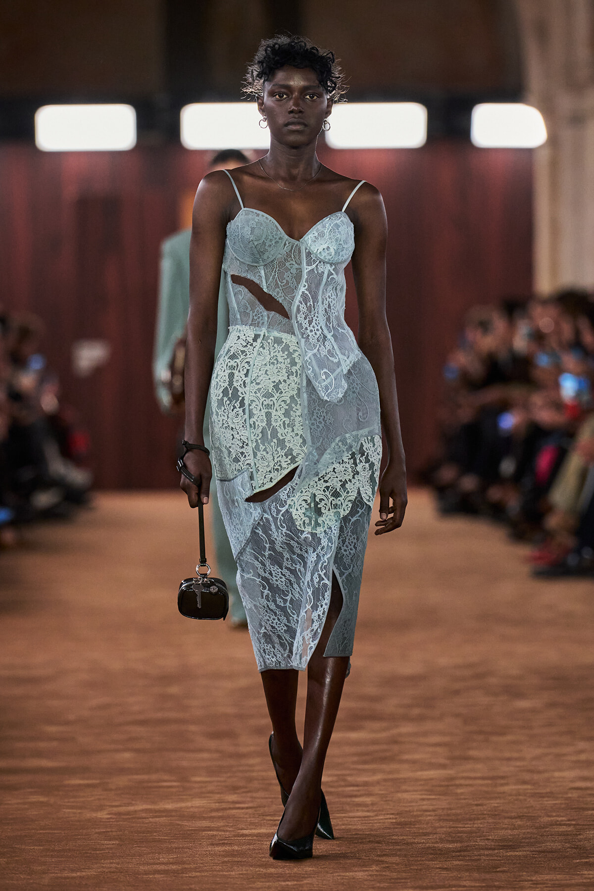 Model walks a runway in a pale blue lace midi dress with cutouts, paired with black heels and a tiny black handbag.