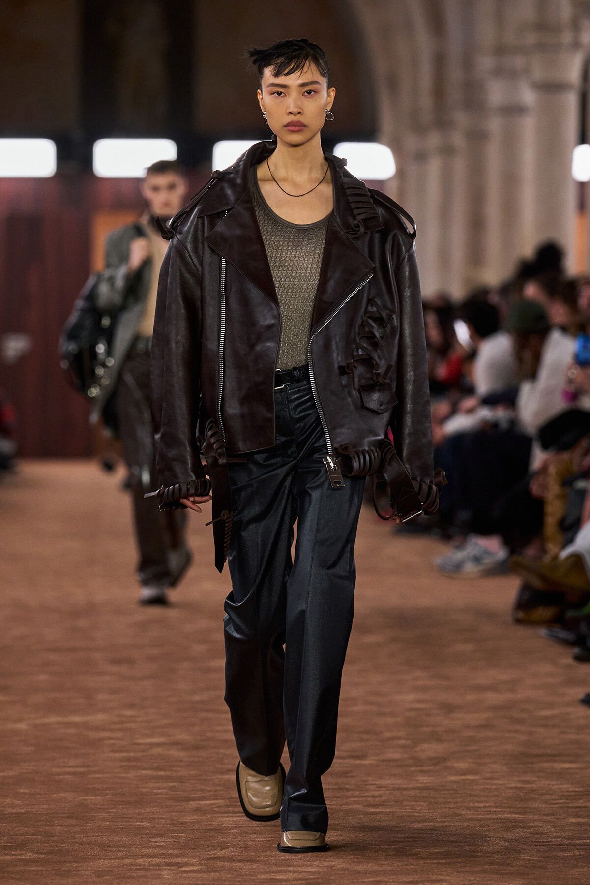 Model walks a runway in an oversized black leather jacket over an olive knit top and dark trousers, with a neutral expression as the audience watches.