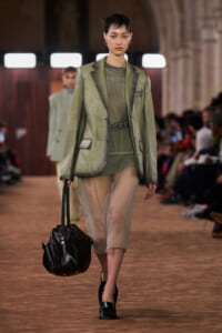 Model walking a runway in an olive-green blazer and sheer green dress, carrying a large black handbag.