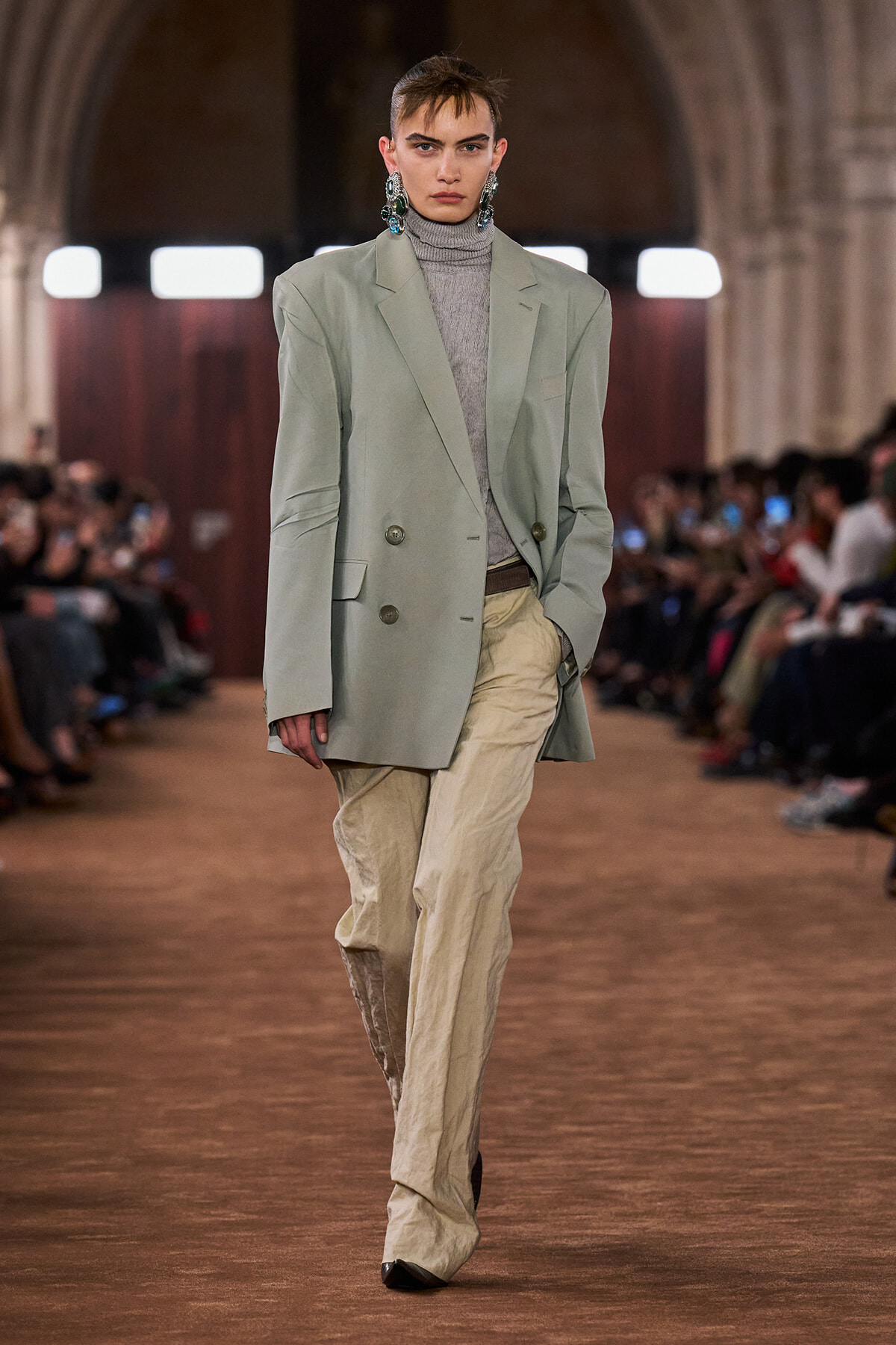 Model walking a runway in an oversized sage blazer, grey turtleneck, and crinkled beige trousers; bold earrings stand out.