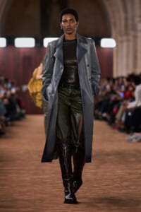 Model walks the runway in a long gray leather trench over a black top and dark green leather pants with tall boots. Audience blurred in background.
