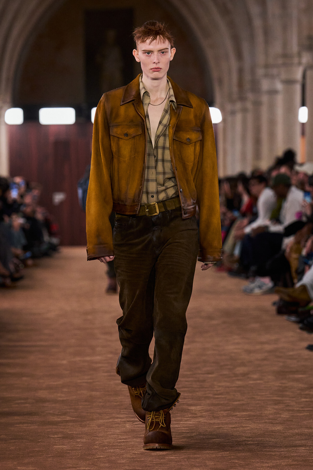 Male model walks a runway in a brown leather jacket over a plaid shirt and dark brown pants, wearing boots in earthy tones.