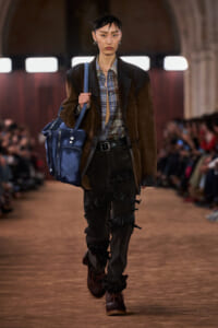 Model walking a runway carrying a large blue bag over her shoulder, wearing a brown blazer, plaid shirt, and distressed dark pants with buckled details.