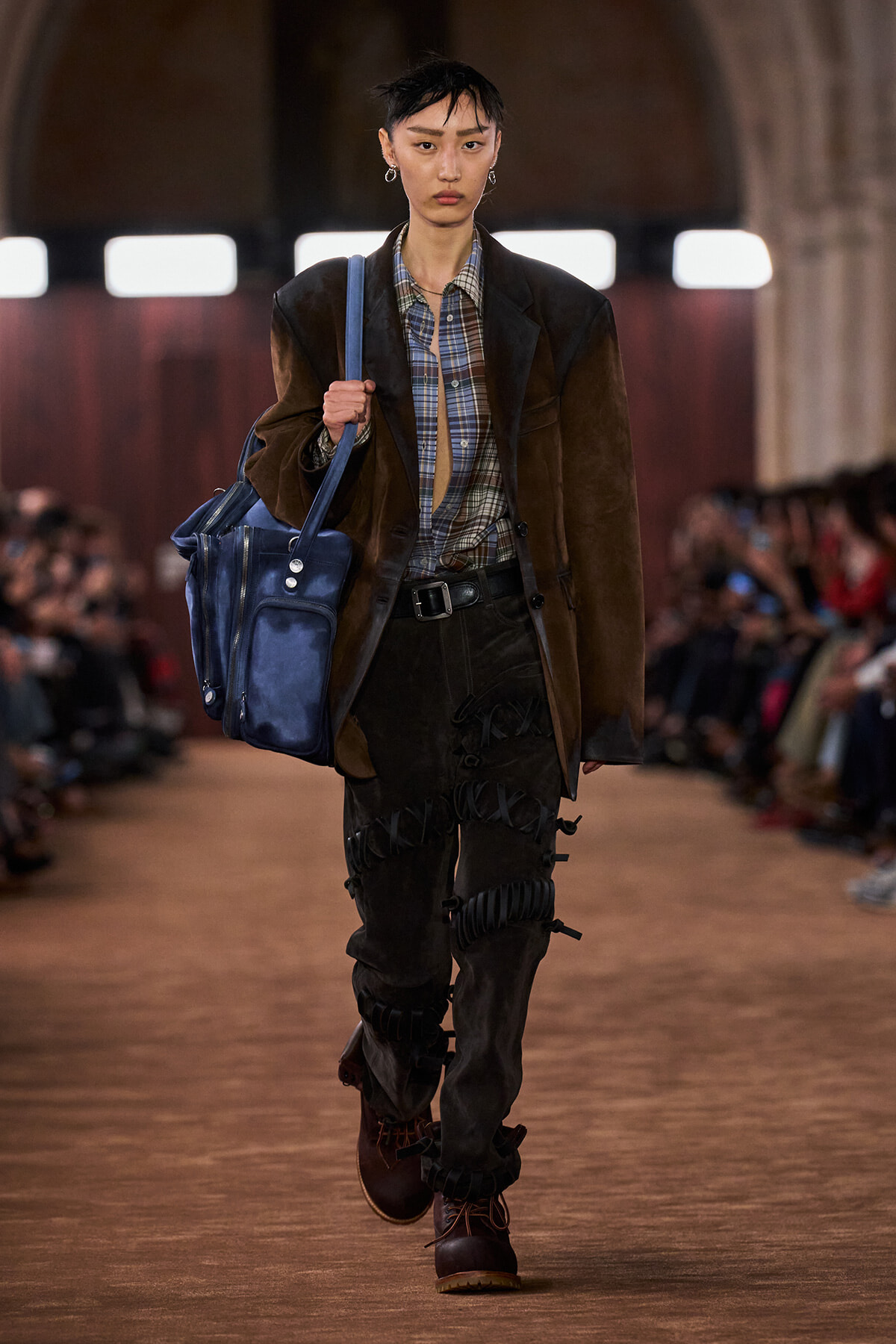 Model walking a runway carrying a large blue bag over her shoulder, wearing a brown blazer, plaid shirt, and distressed dark pants with buckled details.