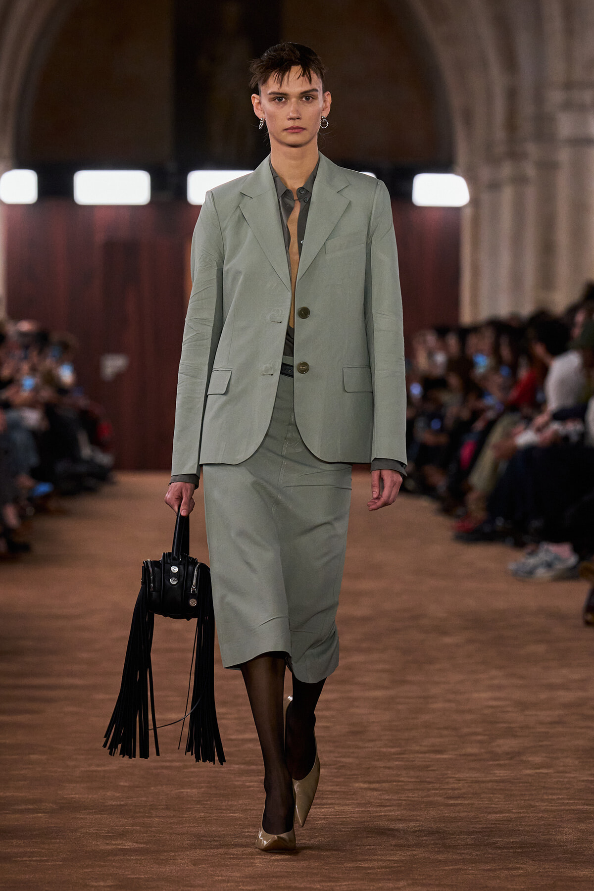Model walking a runway in a sage-green tailored suit with a matching skirt and beige heels, carrying a black fringed handbag.