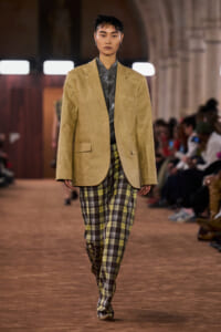 Model walks a runway wearing an oversized tan blazer over a metallic gray shirt and yellow-green plaid trousers, with audience in the background