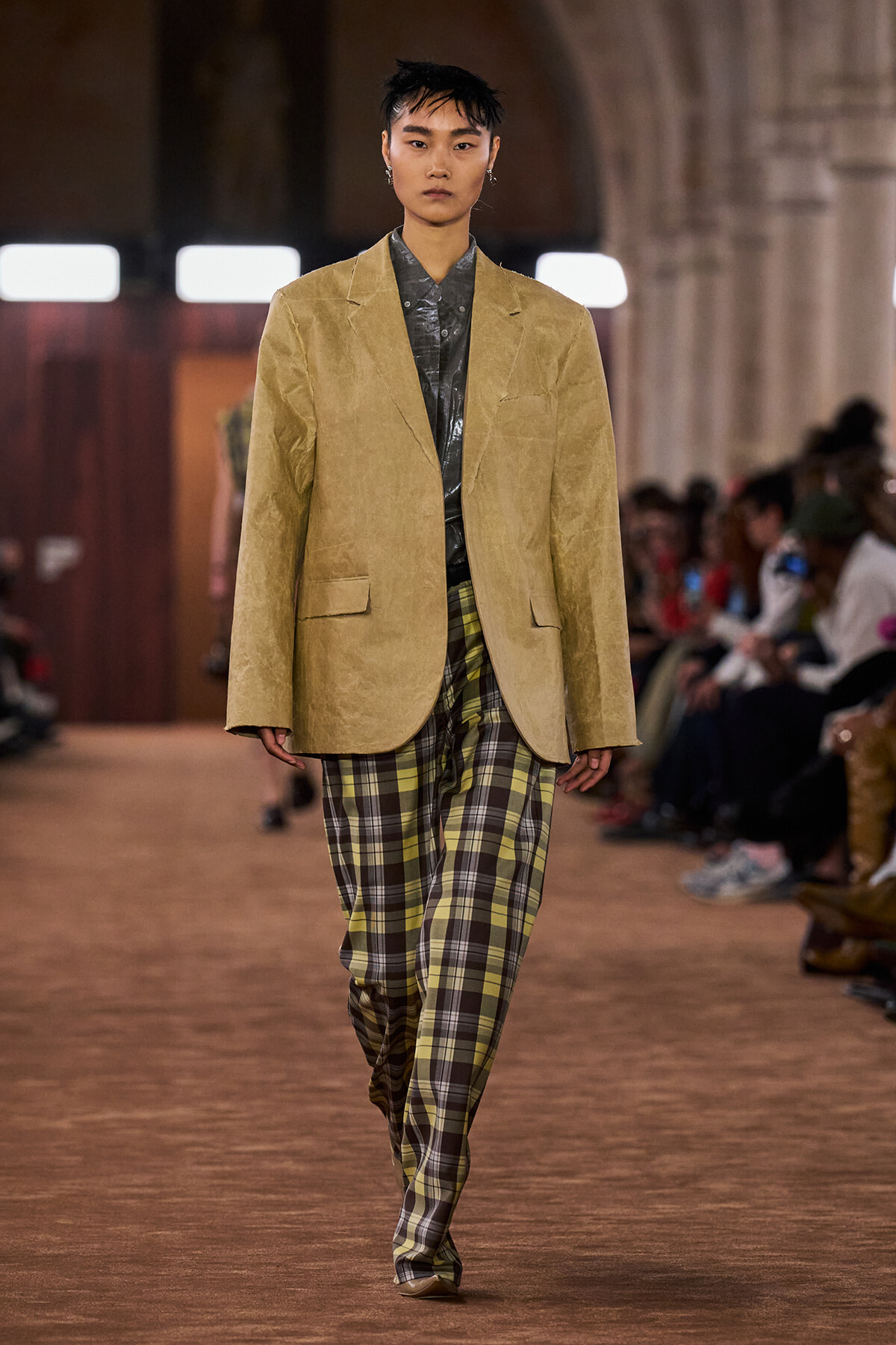 Model walks a runway wearing an oversized tan blazer over a metallic gray shirt and yellow-green plaid trousers, with audience in the background