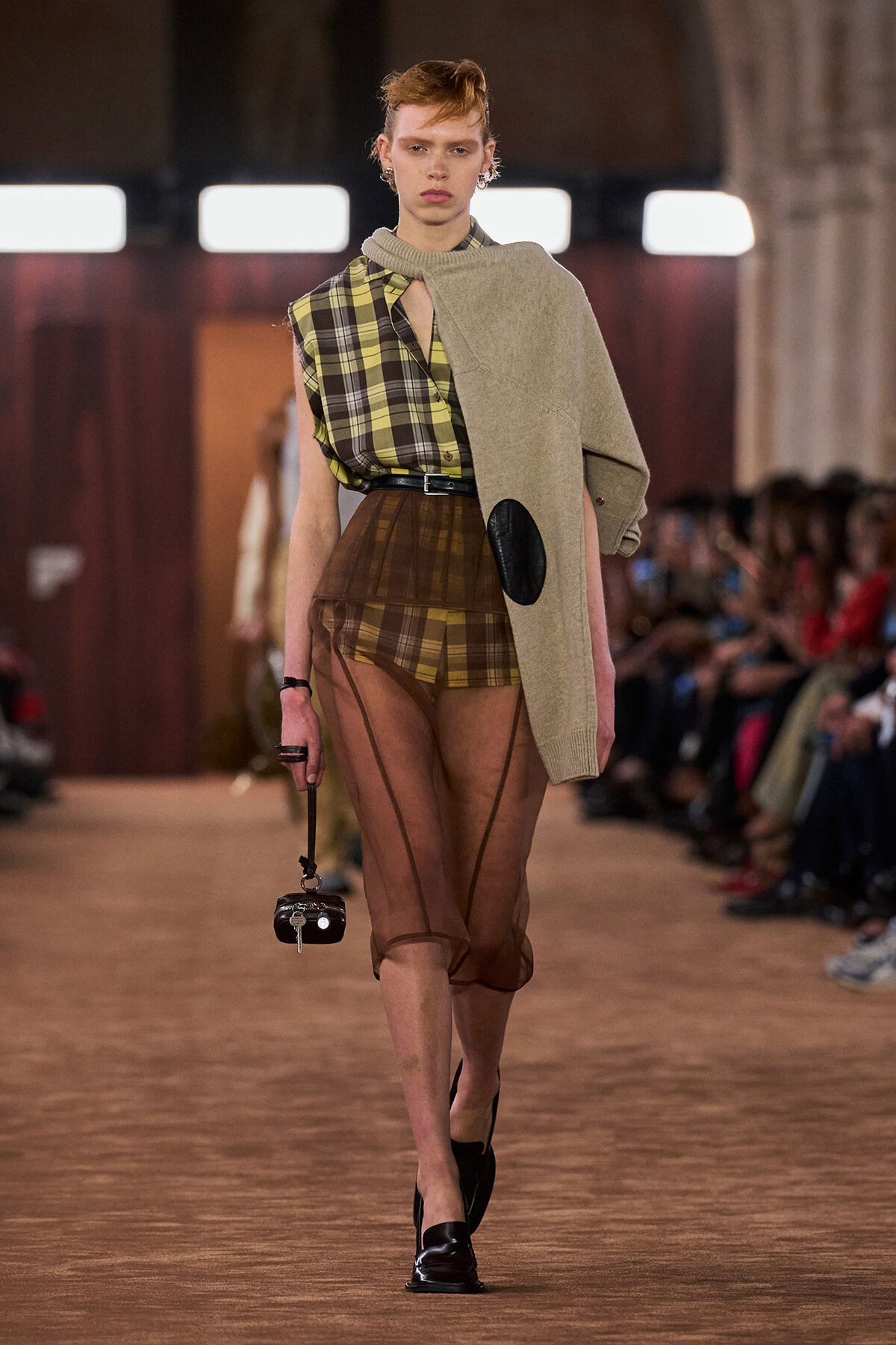 Runway model wearing a green-yellow plaid blouse with brown plaid shorts, sheer brown tights, and a beige scarf draped over one shoulder, carrying a small black handbag.