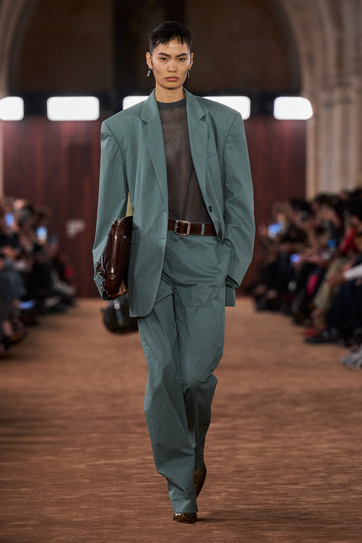 Model on a runway wearing an oversized teal suit with a sheer black top, brown belt, and brown briefcase-style bag draped over the arm.