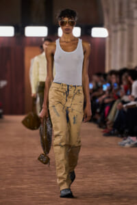 Model walks a runway wearing a white sleeveless top, distressed tan cargo pants, brown sunglasses, and a studded handbag.