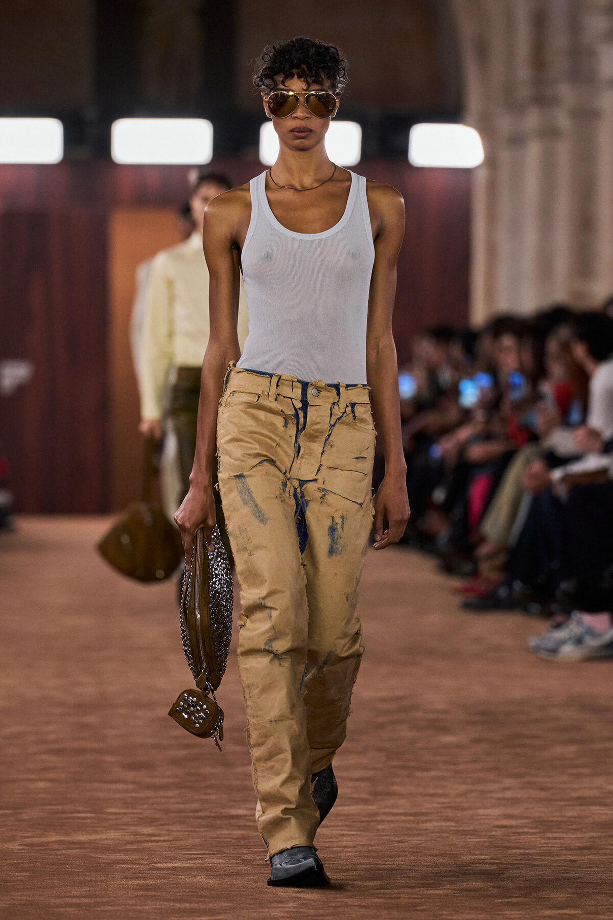Model walks a runway wearing a white sleeveless top, distressed tan cargo pants, brown sunglasses, and a studded handbag.