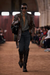 Model walks a fashion show wearing a brown leather blazer, open green shirt, dark pants, and tall cowboy boots while carrying a black handbag and wearing sunglasses on a catwalk.