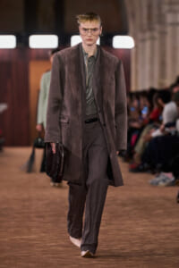 Model on a runway wearing an oversized brown coat over a gray-green shirt and matching trousers, holding a dark satchel.