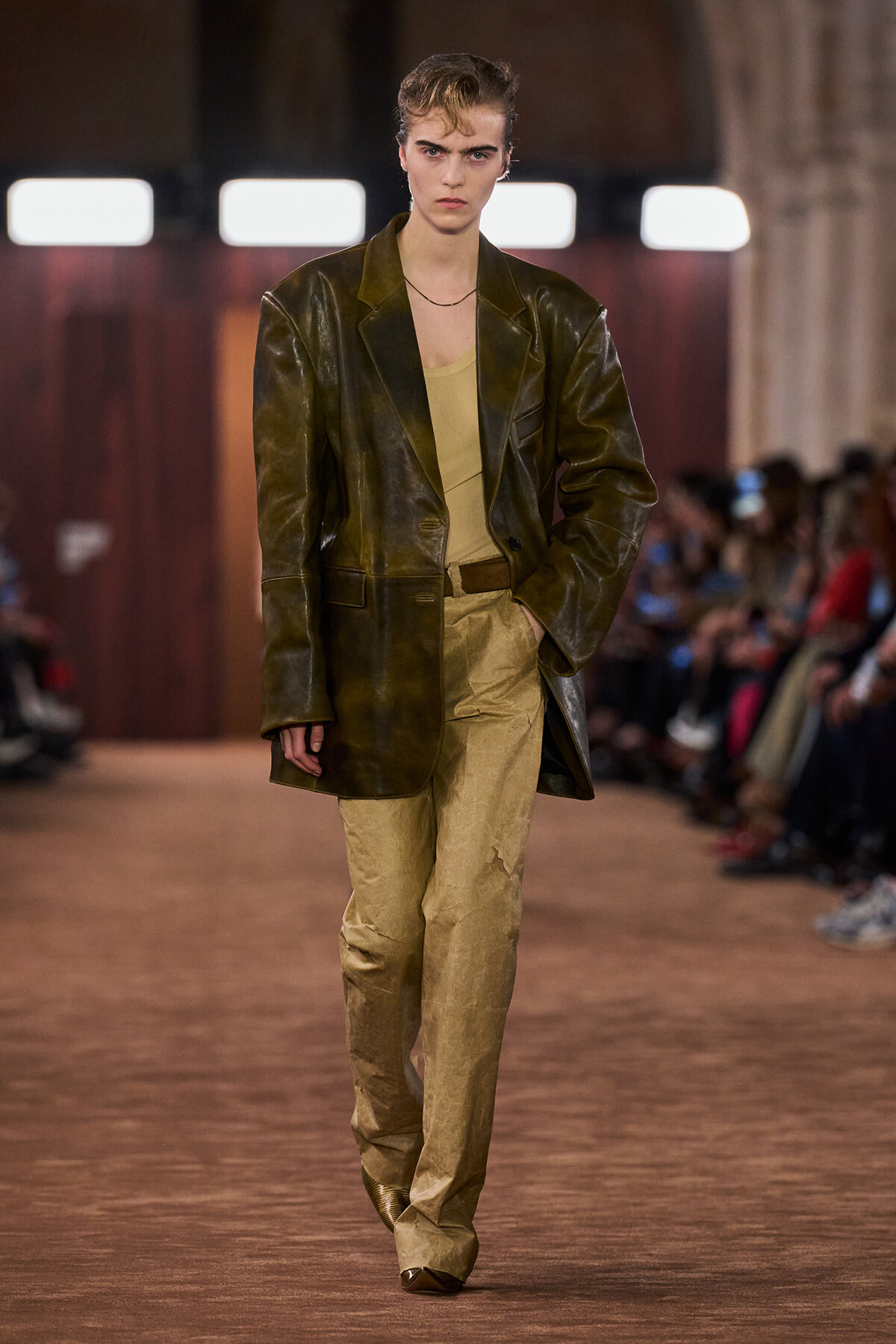 Model walking a runway in an oversized olive leather blazer over a beige top and tan pants.