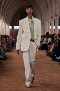 Model walking a fashion runway in an oversized beige suit over a pale green shirt, belt, and loafers in a vaulted venue.