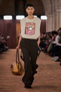 Model on runway wearing a sleeveless beige top with a pink square graphic, olive wide-leg trousers, and carrying a wood-toned handbag.