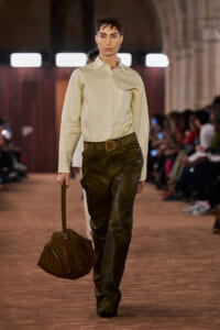 Model walks a runway in a cream button-down shirt, brown belt, and dark leather pants, carrying a brown suede duffel bag with a top handle.