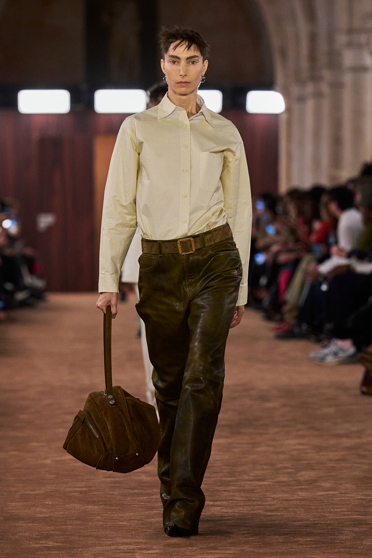 Model walks a runway in a cream button-down shirt, brown belt, and dark leather pants, carrying a brown suede duffel bag with a top handle.