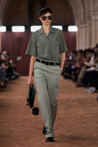 Model walking a runway in olive utility shirt and matching trousers, black belt, sunglasses, and a black clutch bag.