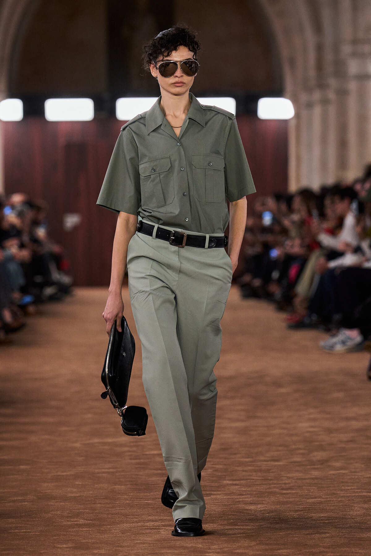 Model walking a runway in olive utility shirt and matching trousers, black belt, sunglasses, and a black clutch bag.