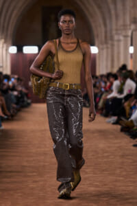 Model walking a runway in a mustard tank top, metallic gray wide-leg pants, and a tan belt, carrying a studded brown bag over the shoulder.