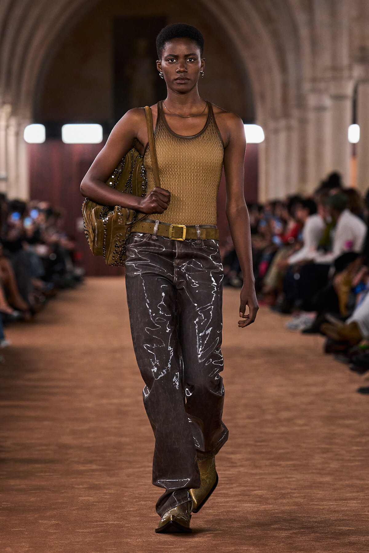 Model walking a runway in a mustard tank top, metallic gray wide-leg pants, and a tan belt, carrying a studded brown bag over the shoulder.