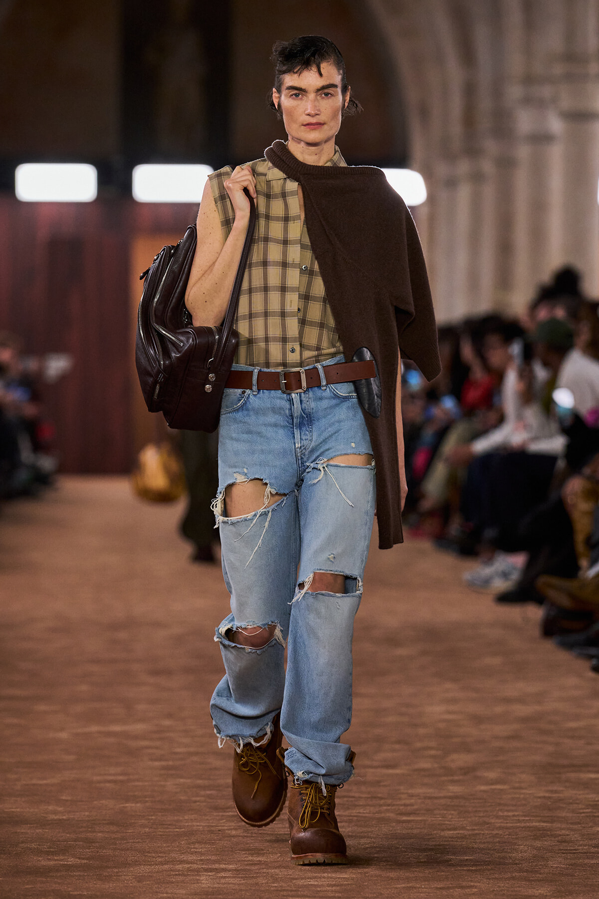 Model walking a runway wearing a plaid button-up tucked into ripped light-wash jeans with a brown belt, brown boots, and a dark brown cape draped over one shoulder, carrying a large black backpack