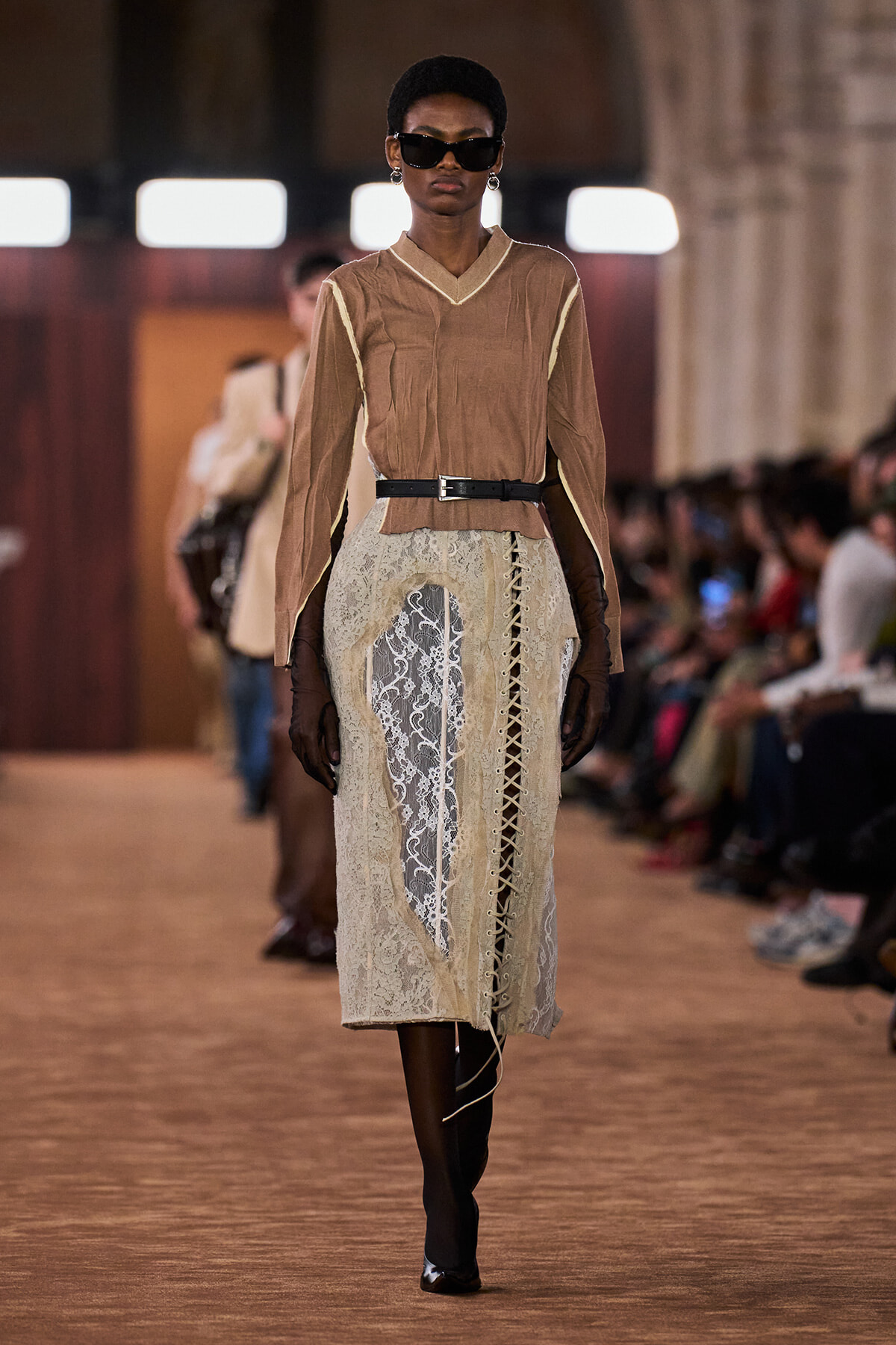 Model walking a runway in a taupe blouse with contrasting cream trim, a lace pencil skirt, and black belt, accessorized with dark sunglasses and gloves.