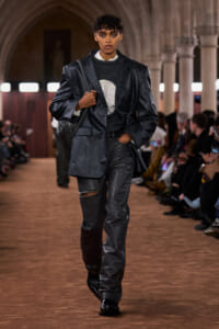 Runway model in an oversized black leather blazer and matching pants, carrying a shoulder bag as they walk the catwalk.