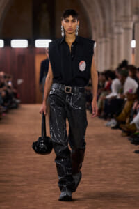 Model walks a fashion runway in a sleeveless black shirt with a circular pocket patch, black marble-pattern pants, and a belt, carrying a black bag.