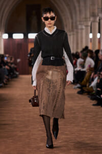 Model walking runway in oversized black V-neck top over white shirt, taupe lace pencil skirt, black belt, sunglasses, and heeled shoes, carrying a small brown handbag.