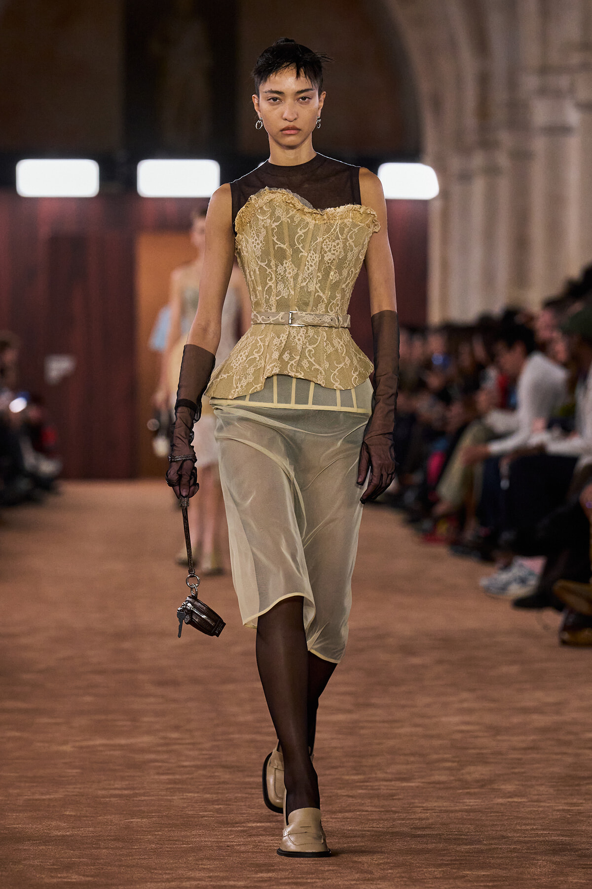 Model on a runway wearing a beige lace corset over a sheer black top, belted waist, pale satin skirt, brown gloves, and beige shoes.