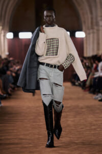 Model walks a runway wearing a cream oversized sweater with a plaid chest patch over a green plaid shirt, ripped light denim jeans, and knee-high boots, carrying a dark coat over the shoulder.