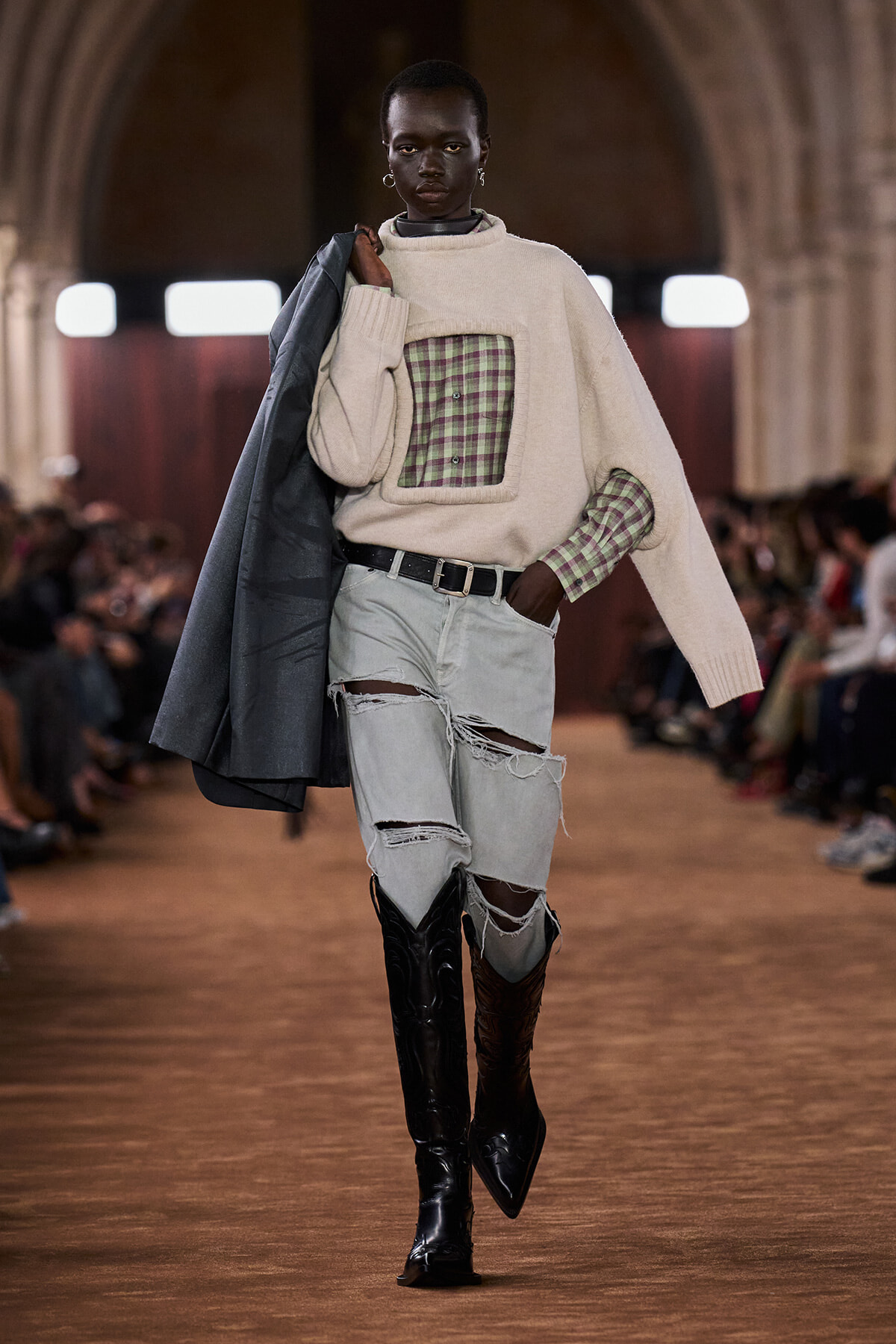 Model walks a runway wearing a cream oversized sweater with a plaid chest patch over a green plaid shirt, ripped light denim jeans, and knee-high boots, carrying a dark coat over the shoulder.