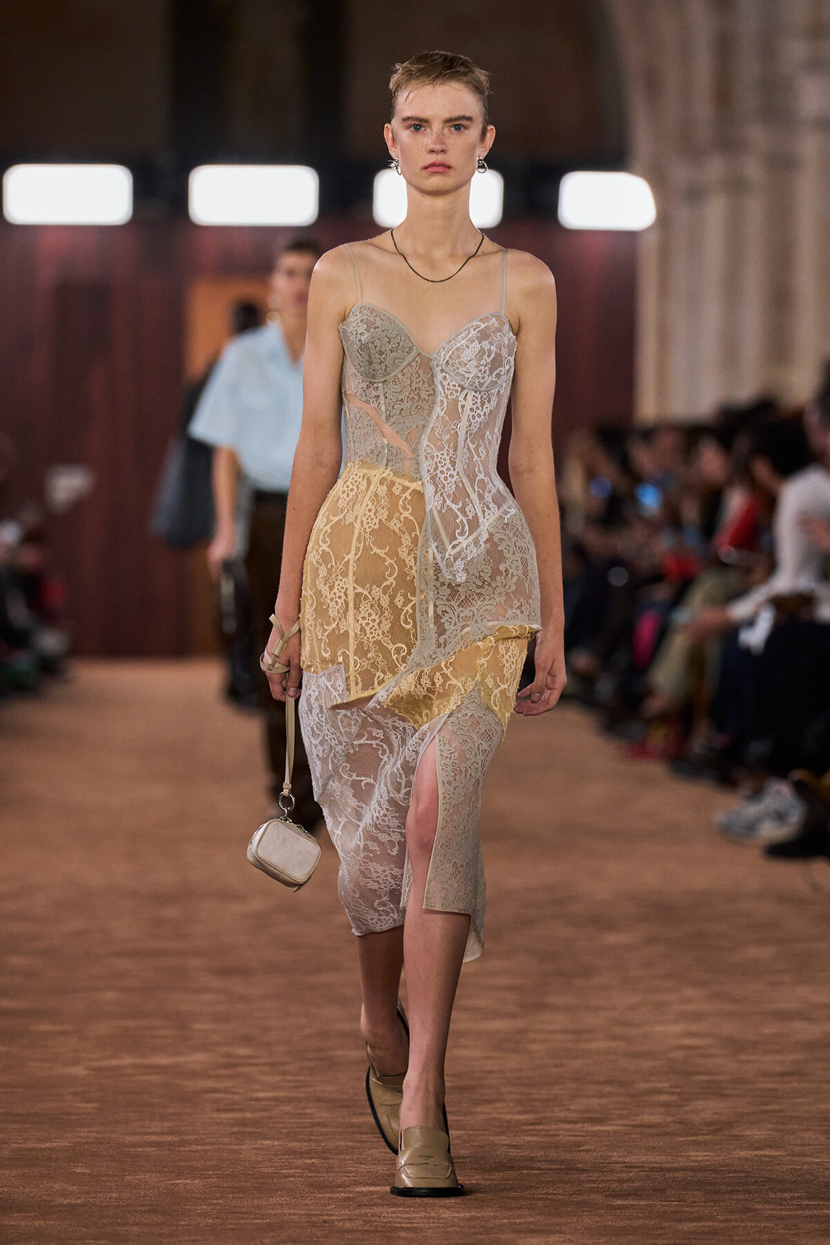 Model walking a runway in a sheer lace midi dress in cream and beige tones, carrying a small cream purse.