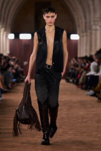 Model walking a runway in a black sleeveless leather vest, open at the chest, with black pants, tall black boots, and a large fringe leather bag in hand.