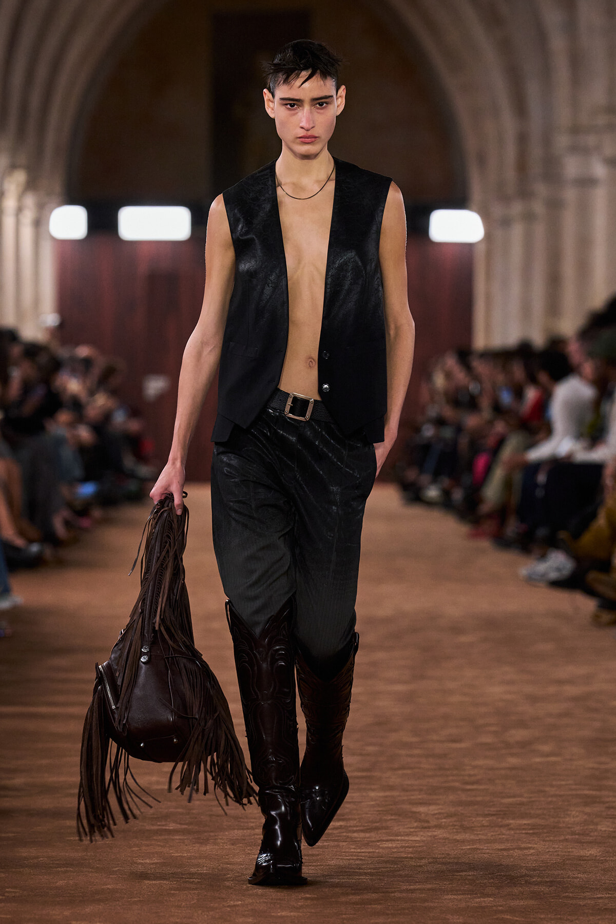 Model walking a runway in a black sleeveless leather vest, open at the chest, with black pants, tall black boots, and a large fringe leather bag in hand.
